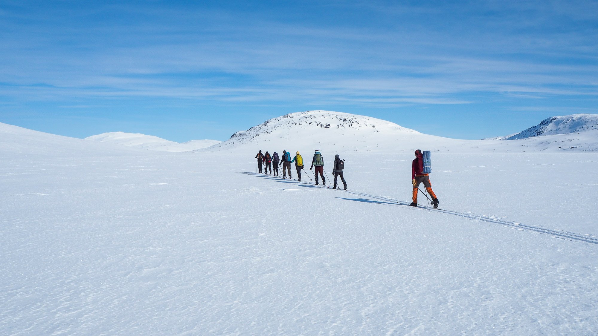 Joukko retkeilijöitä hiihtää jonossa kohti lumisia tuntureita. 